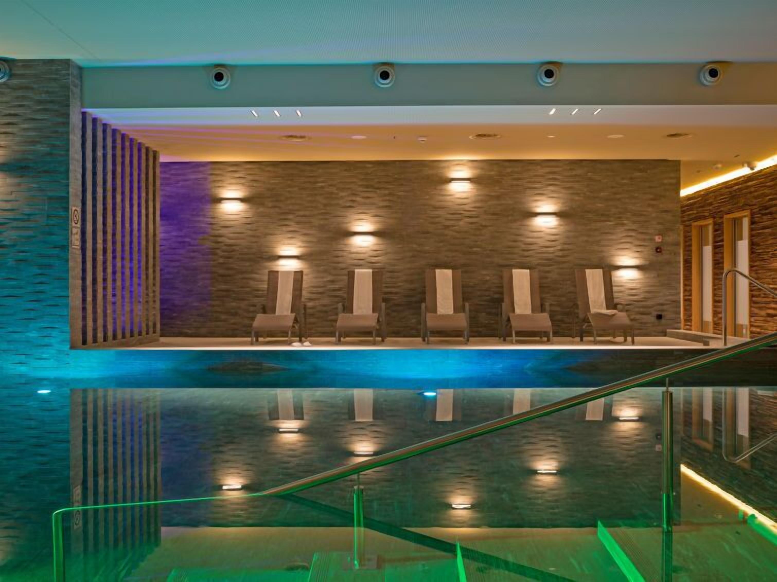 A modern indoor swimming pool with a glass barrier, multiple lounge chairs, and a stone-textured wall.