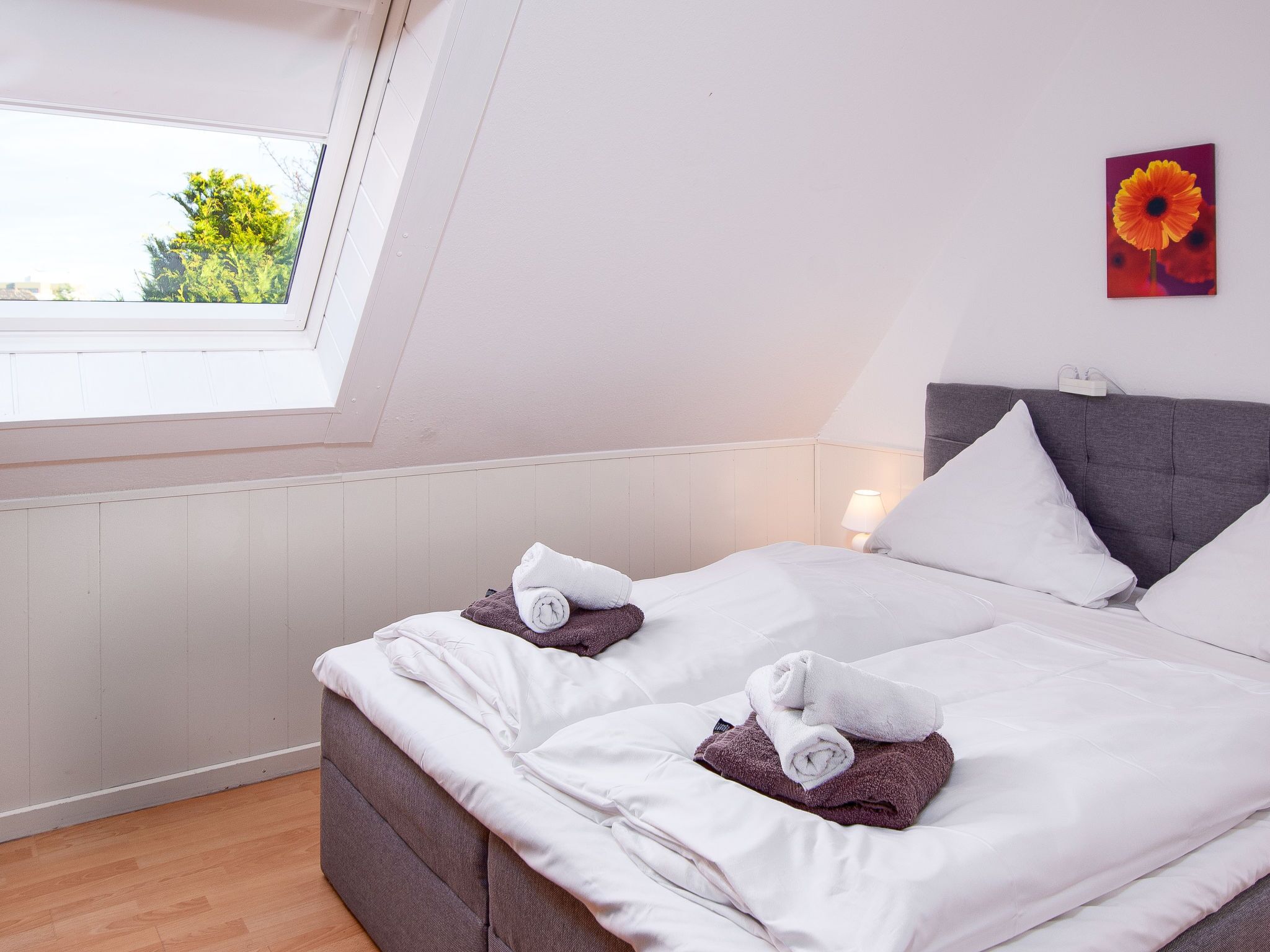 A neatly made bed with white and purple towels, a grey headboard, and a window with a view of greenery.