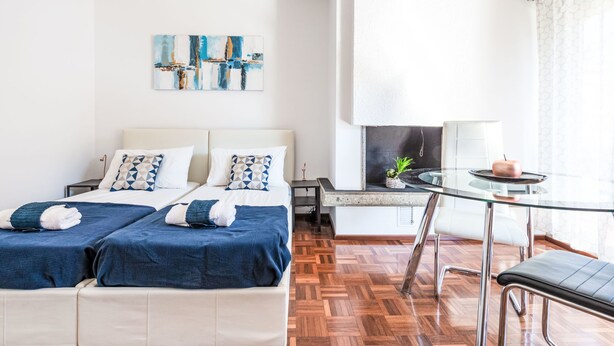 Moderni hotellihuone, jossa on kaksi sänkyä, pieni ruokailutila lasipöydällä sekä suuri ikkuna, jossa on verhot.