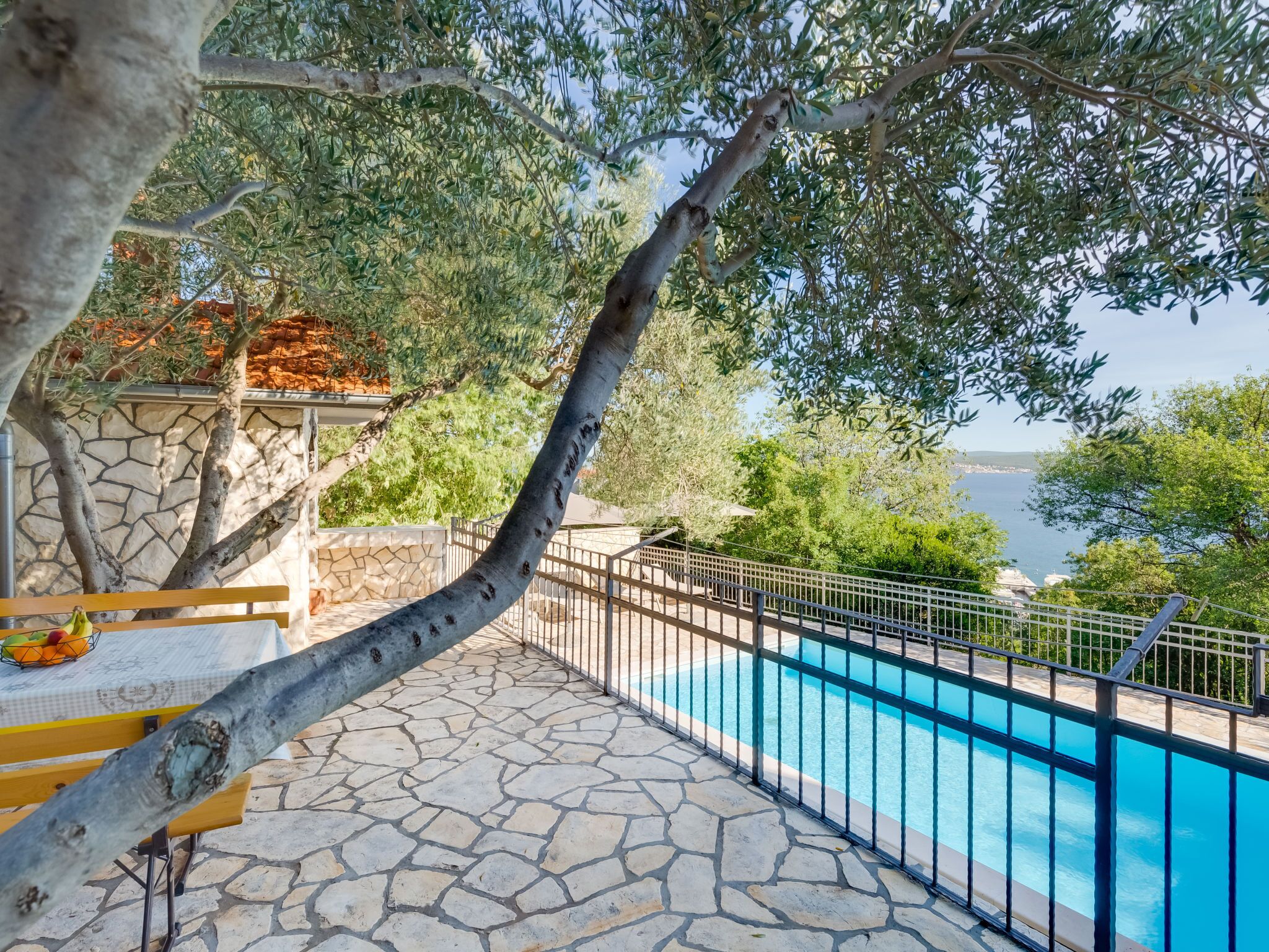 A stone-paved pool area with a metal fence, surrounded by trees and a view of the sea.