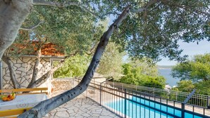 Area piscina con pavimentazione in pietra, recinzione in metallo, alberi e vista sul mare.