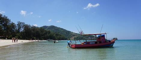 A beach with a red boat anchored near the shore, clear turquoise water, and a forested hill in the background.