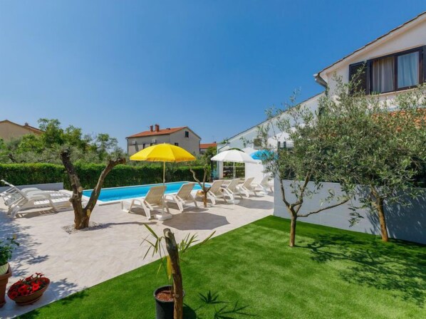 A pool area with lounge chairs and umbrellas, surrounded by greenery and a building. - Suncana by Interhome (Porec)