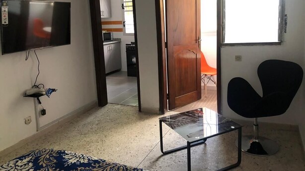 Ein Zimmer mit an der Wand montiertem Fernseher, einem kleinen Tisch, einem schwarzen Stuhl und einer Tür zu einer Küchenzeile.