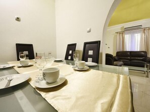 Un coin repas avec une table en verre dressée pour quatre personnes, comprenant des assiettes, des bols et des verres à vin. On y trouve deux chaises noires avec des coussins d’assise blancs. Un canapé en cuir est visible au fond.