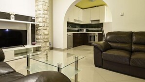 A modern living room with a glass coffee table, leather sofa, and a flat-screen TV. A stone accent wall and an arched doorway lead to a kitchen with dark cabinets and a microwave. - Apartment Anić by Interhome (Novigrad (Zadar))
