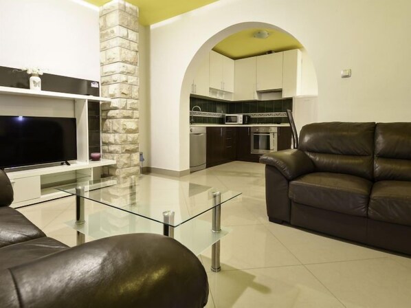 A modern living room with a glass coffee table, leather sofa, and a flat-screen TV. A stone accent wall and an arched doorway lead to a kitchen with dark cabinets and a microwave.