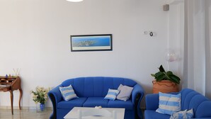 A modern living room with a blue sofa, white table, and chairs. There is a ceiling light, a framed picture on the wall, and a potted plant.
