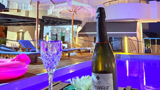 A bottle of Prosecco and a glass on a wooden table with a pool and lounge chairs in the background.