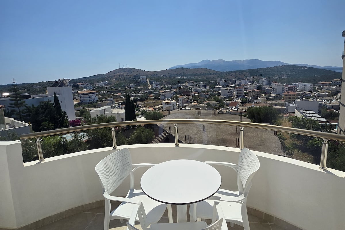 A balcony with a white table and chairs, overlooking a cityscape with buildings and mountains.