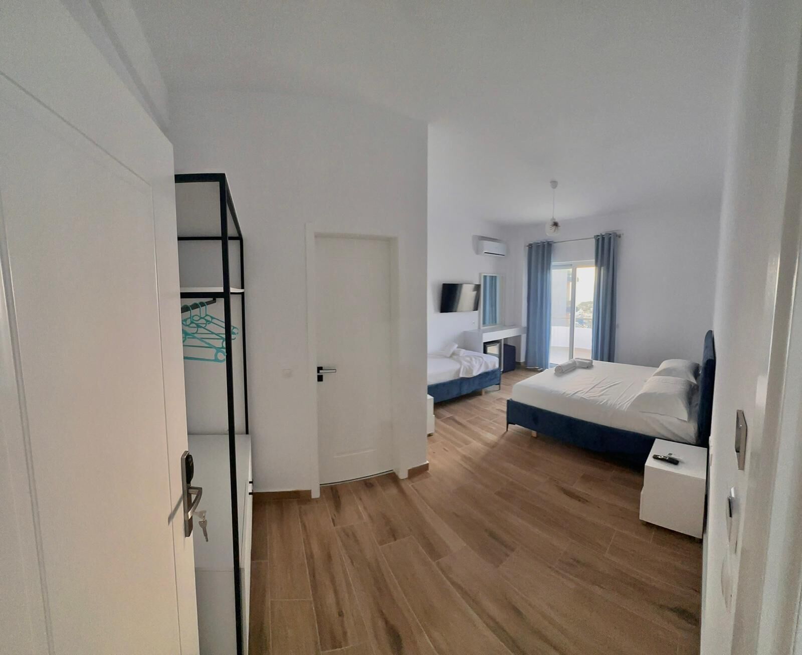 Quarto de hotel com piso de madeira, uma cama, escrivaninha, cadeira, televisão e varanda com cortinas.
