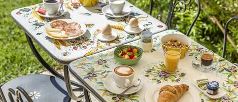 Una mesa colocada al aire libre con mantel floreado, platos de comida, tazas de café y un croissant.