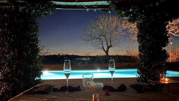 Una mesa de comedor con dos copas de vino, una vela y una botella de champán, con vistas a una piscina y un árbol.