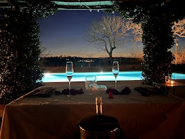 A dining table with two wine glasses, a candle, and a bottle of champagne, overlooking a swimming pool and a tree.