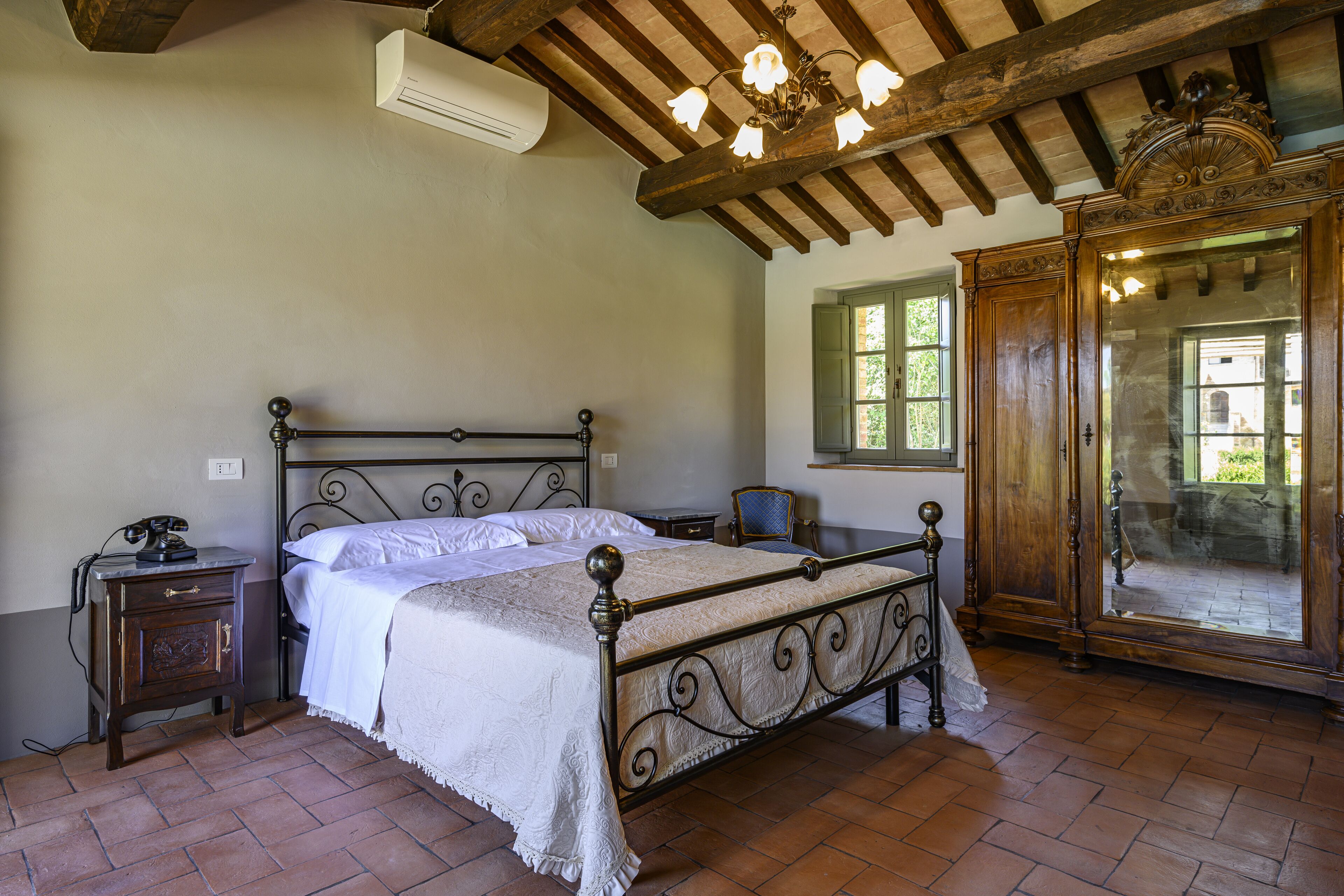 A bedroom with a wooden bed frame, a nightstand with a telephone, a mirror, and a window with green shutters.