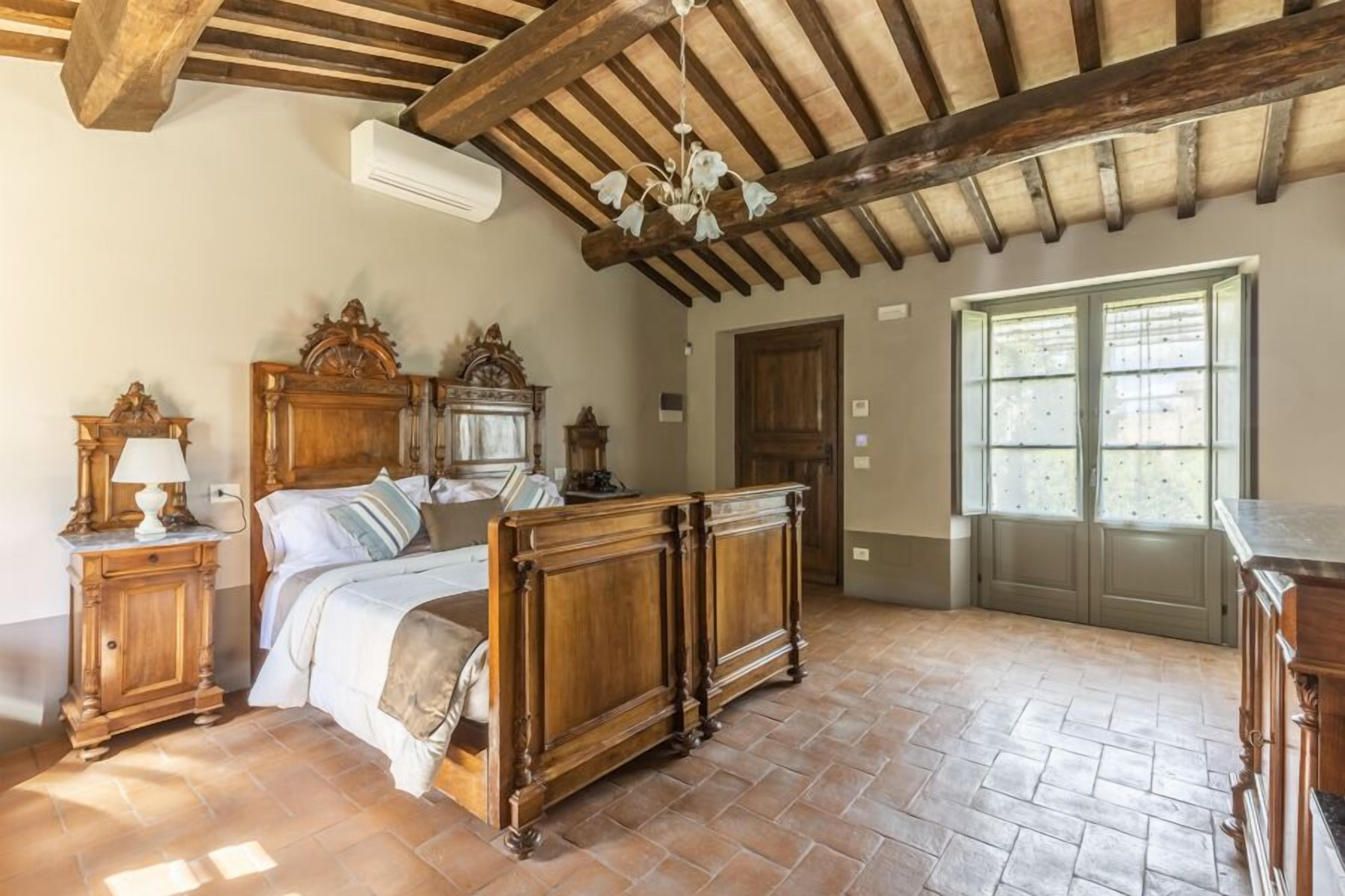 A bedroom with a large bed, wooden headboard, and a chandelier. There is a wooden dresser and a window with blinds.