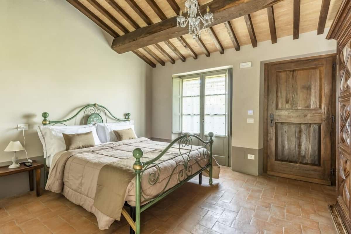 A bedroom with a bed, wooden ceiling beams, a chandelier, a wooden door, and a window with blinds.
