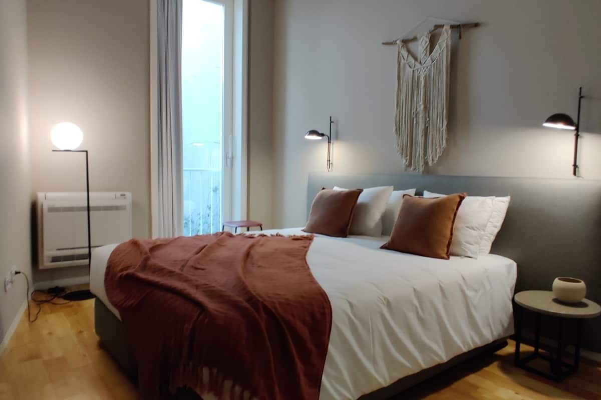 Un dormitorio con cama, mesita de noche, lámpara y un tapiz de macramé en la pared.