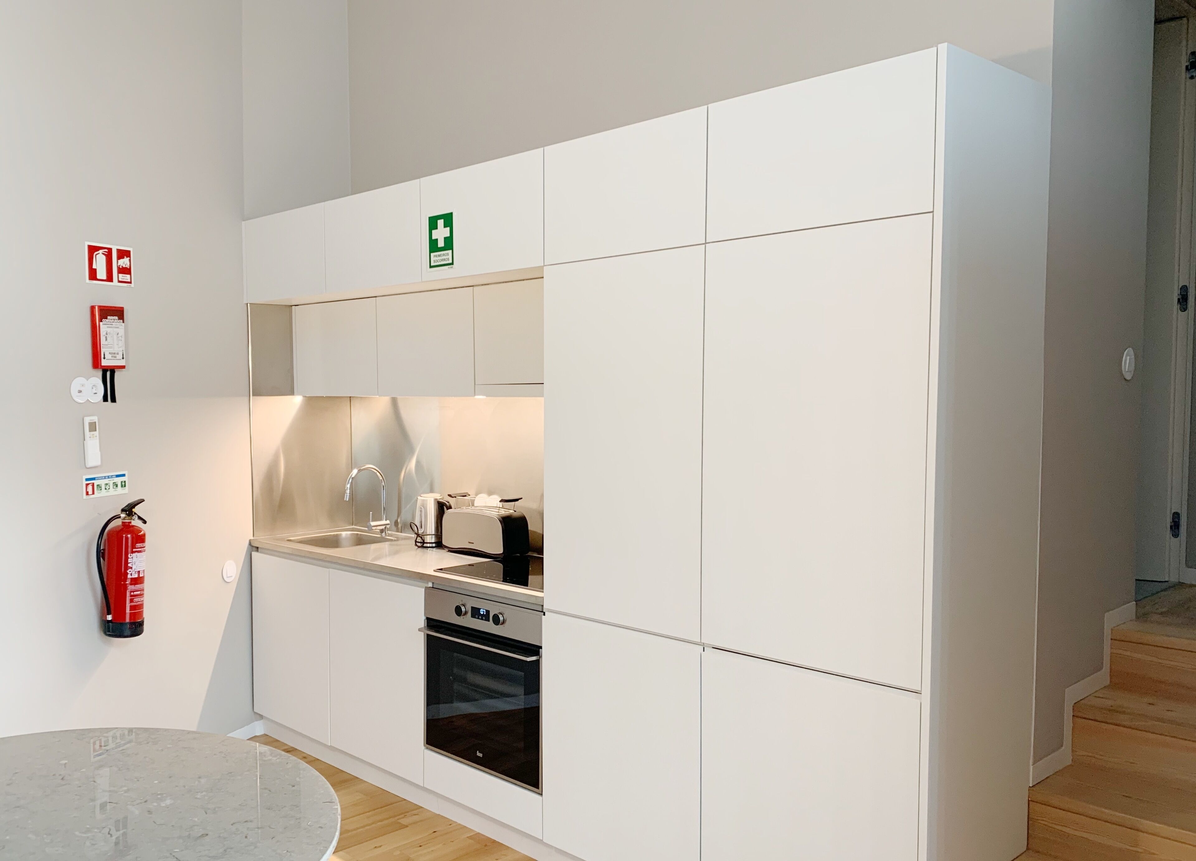 Une cuisine moderne avec des meubles blancs, un four encastré, un évier et un grille-pain. Un extincteur est fixé au mur et une trousse de premiers secours est visible.