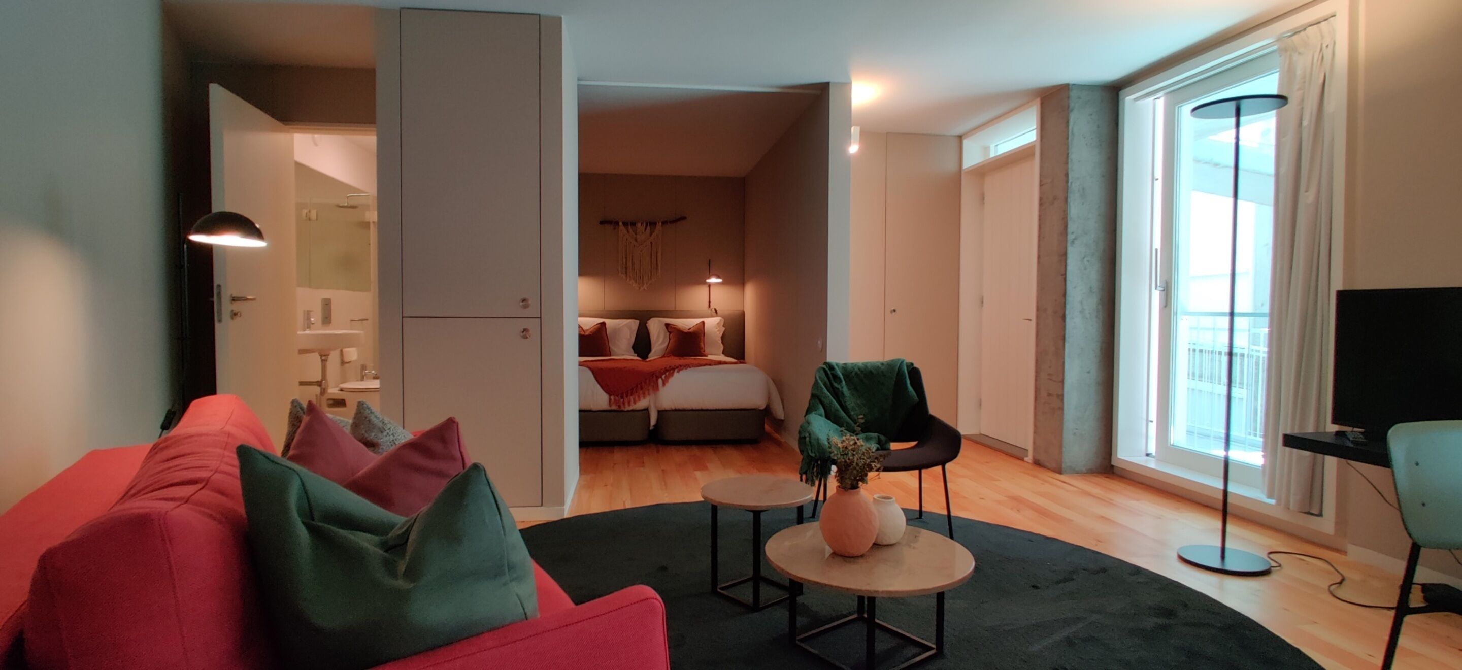 Una sala de estar moderna con un sofá rojo, una mesita de centro pequeña y una silla verde. Se ve una cama con cabecera blanca y una colcha verde al fondo.