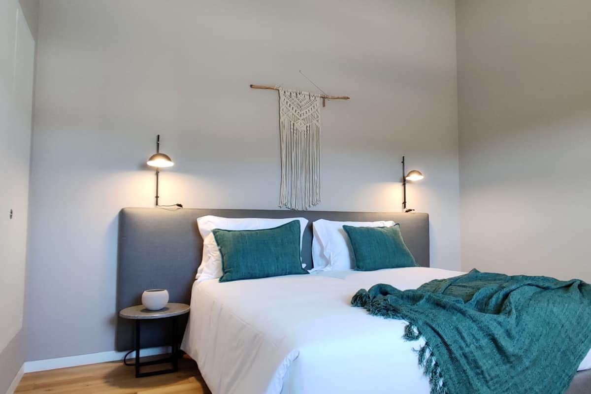 Un dormitorio ordenado con una cama, dos lámparas de noche, un colgador de macramé y una mesita de noche con un cuenco.