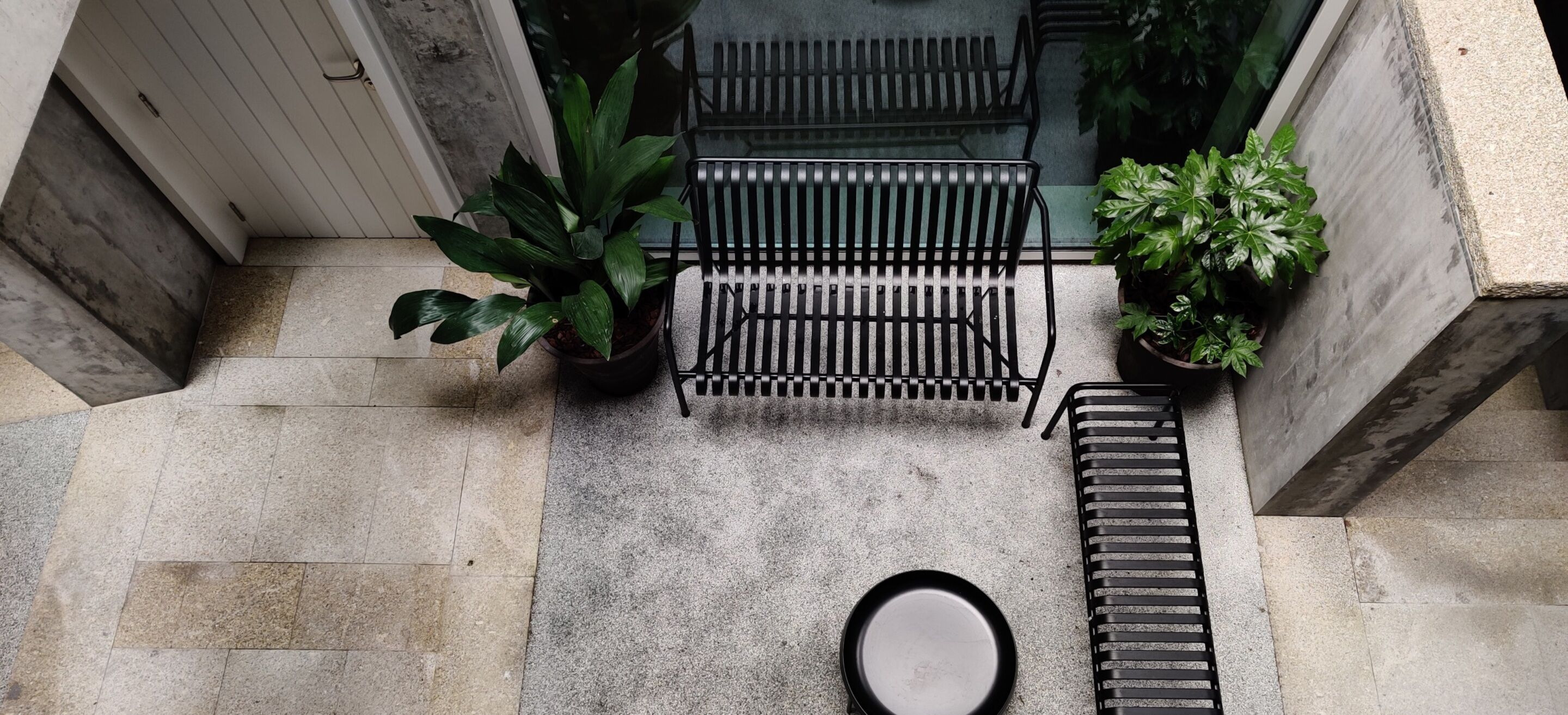 A small outdoor seating area with a metal bench, two potted plants, and a round table.