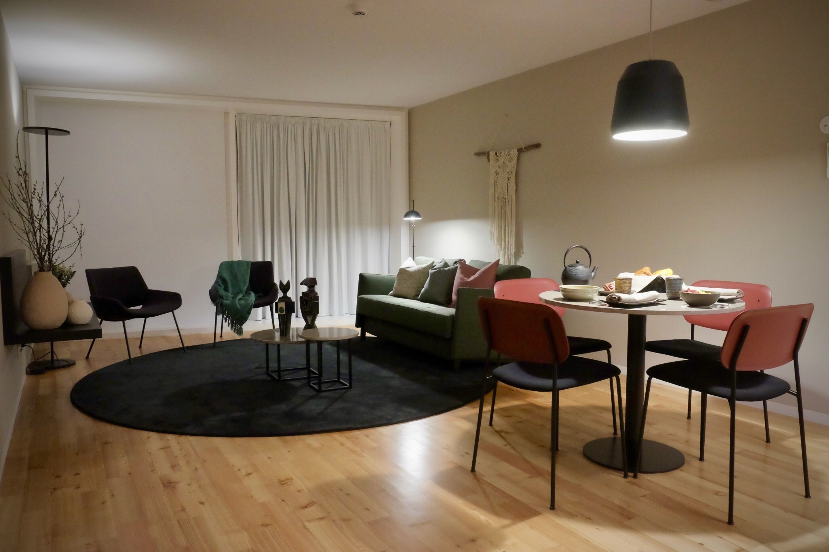 A modern living room with a round dining table, green sofa, and black and red chairs. A black floor lamp and a white curtain are also visible.