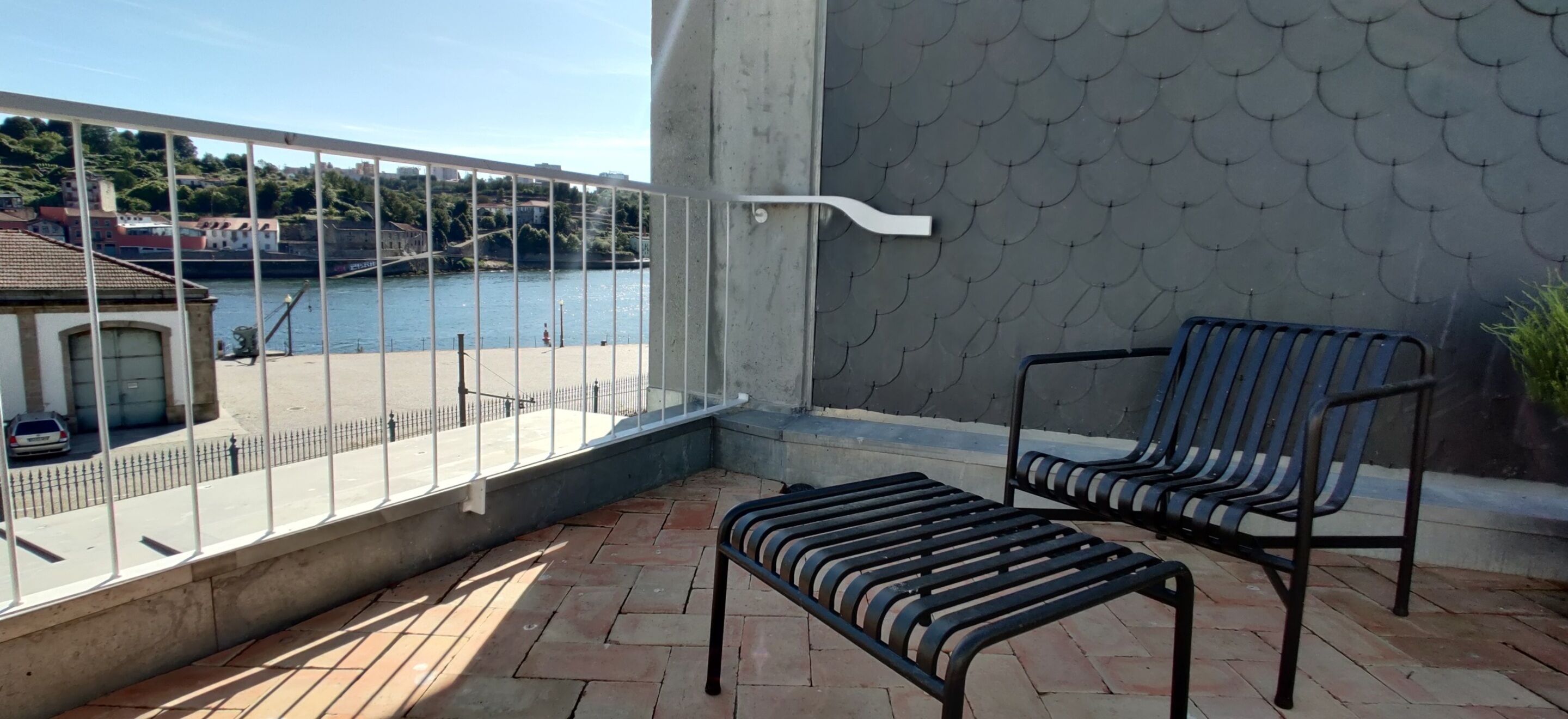 A balcony with two chairs and a small table, overlooking a body of water and a distant town.