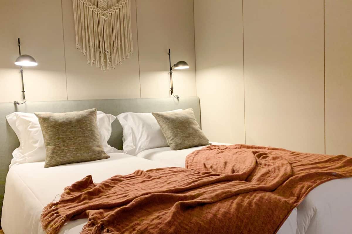 Una cama con ropa de cama blanca y dos almohadas, un edredón marrón y un colgador de macramé en la pared.