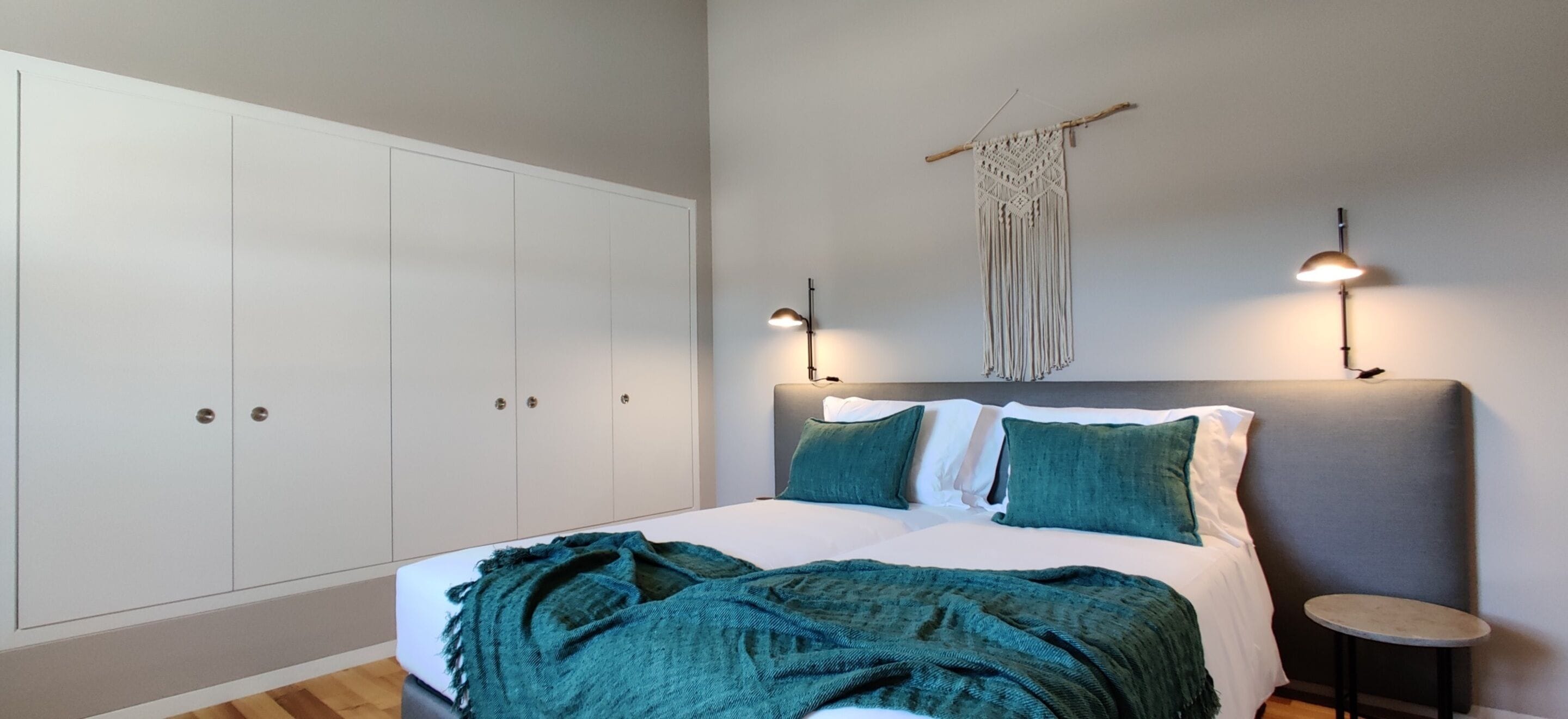 A bedroom with a bed, two bedside lamps, a macrame wall hanging, and a small round table.
