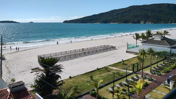 A beach with a clear view of the sea, a sandy walkway, and a building with a sloped roof. - Flat Bellmare (Florianópolis)