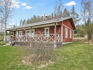 Eine rote Holzhütte mit Veranda und Schornstein, umgeben von Bäumen und Gras.