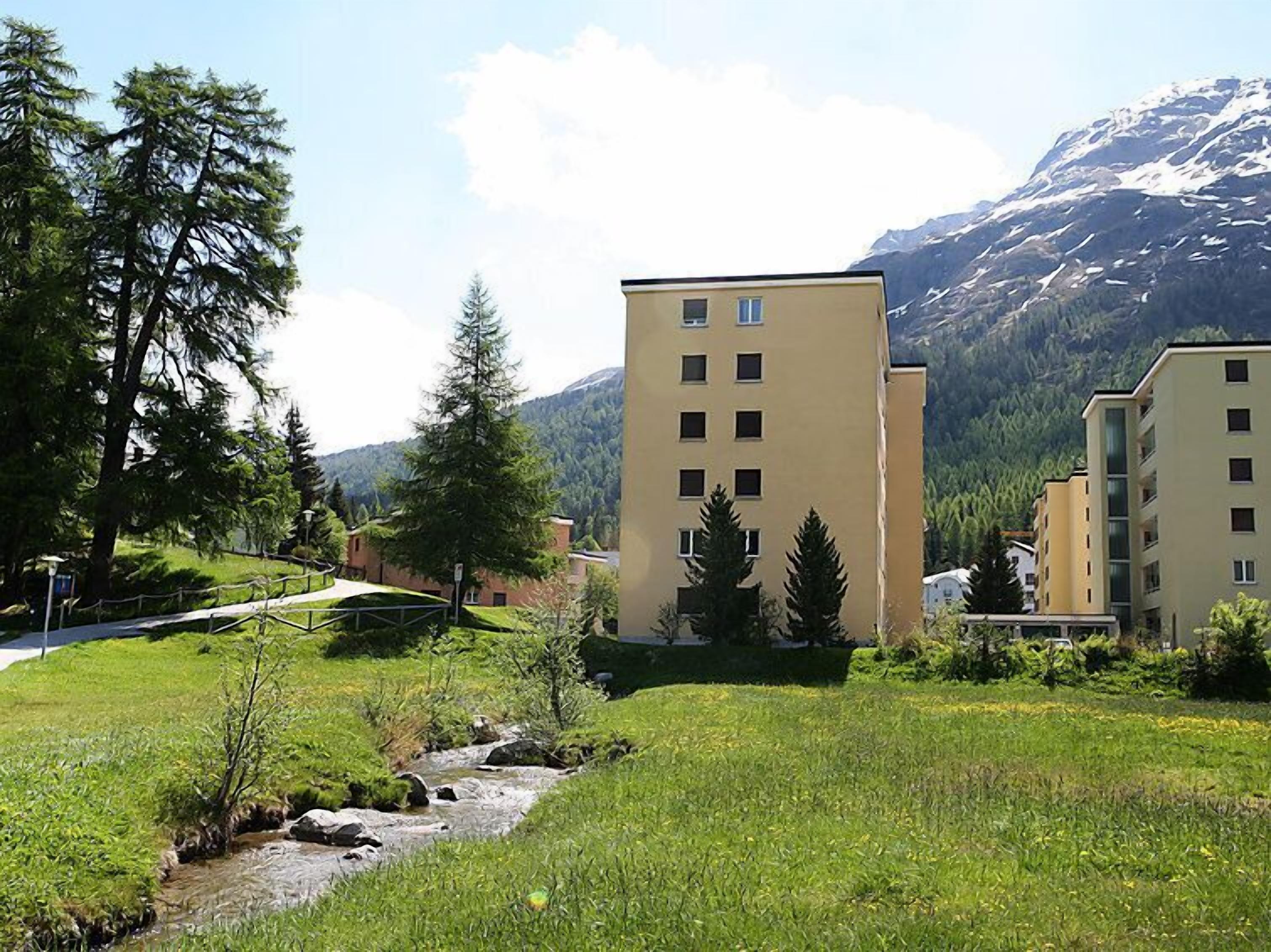 Il s’agit d’un quartier résidentiel traversé par un petit ruisseau, avec de l’herbe verte et des arbres. On y trouve des bâtiments dotés de nombreuses fenêtres, et une montagne se profile à l’arrière-plan.