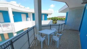 A covered outdoor patio with a table and chairs, a metal railing, and a view of a building with blue and white walls.
