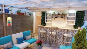 Een terras met een blauwe bank, een kleine tafel met een bloevas, en een bar met stoelen en planken.