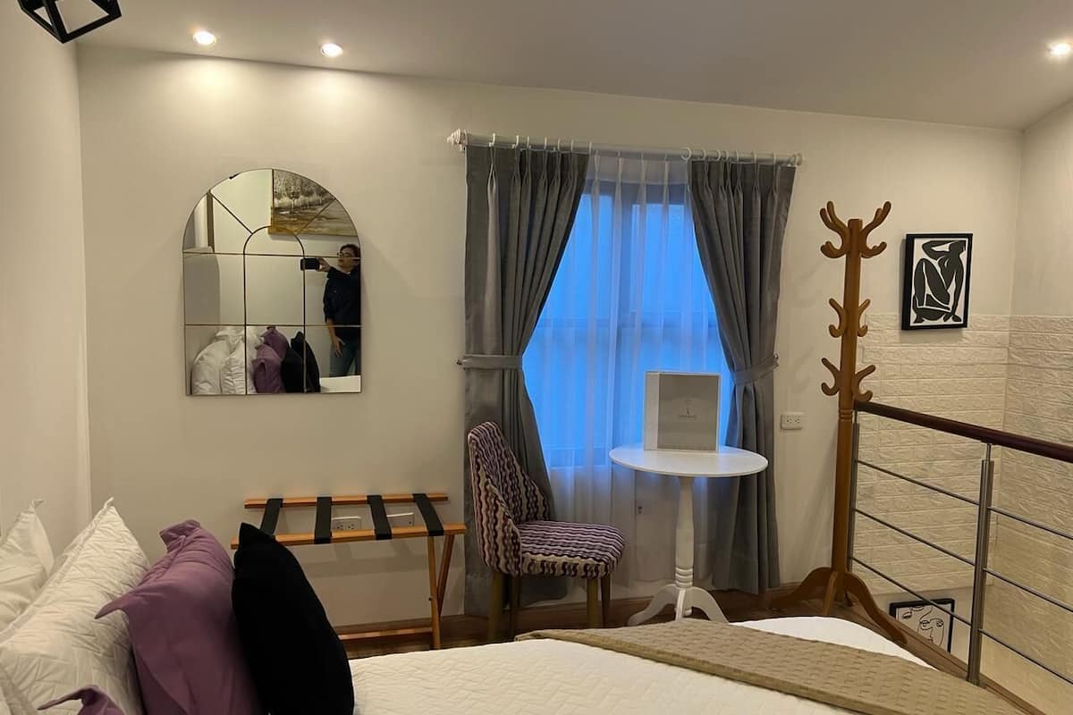 Ett hotellrum med en säng, en stol, ett litet bord, en spegel och ett fönster med gardiner.