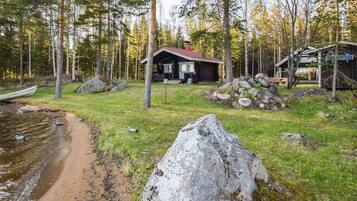 A cabin with a porch, surrounded by a forest, a small dock, and a pile of rocks.