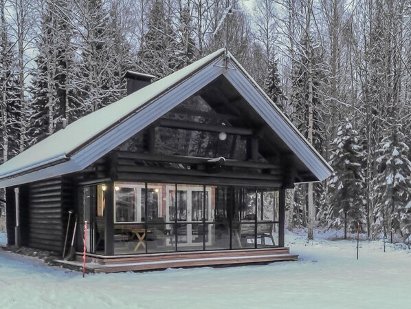A log cabin with a large glass front, surrounded by snow-covered trees.