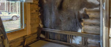 A log cabin interior with a large fur rug, a window with a view of a car outside, and a metal bed frame.