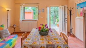 A dining area with a table set for two, a sofa, a radiator, and a view of the garden through the open door. - Contrada Brecciano by Interhome (Città Sant'Angelo)