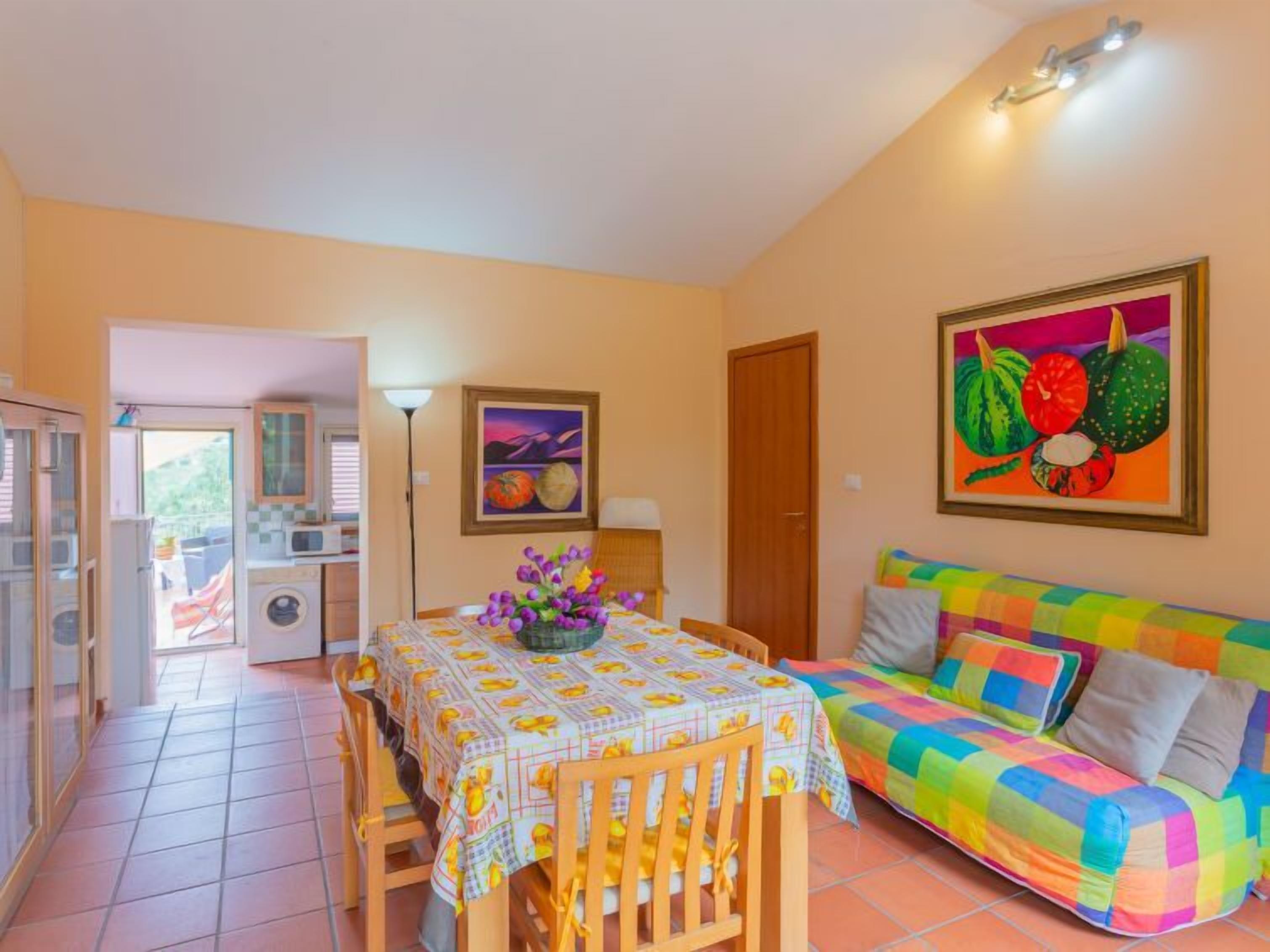 Un espacio de sala de estar acogedor con un sofá colorido, una mesa de comedor con mantel estampado, y una cocina al fondo.