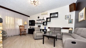 Ein Wohnzimmer mit einer Couch, Sesseln, einem Couchtisch und einem Kamin. Es gibt einen Essbereich mit Tisch und Stühlen sowie eine Küche mit Herd und Schränken.