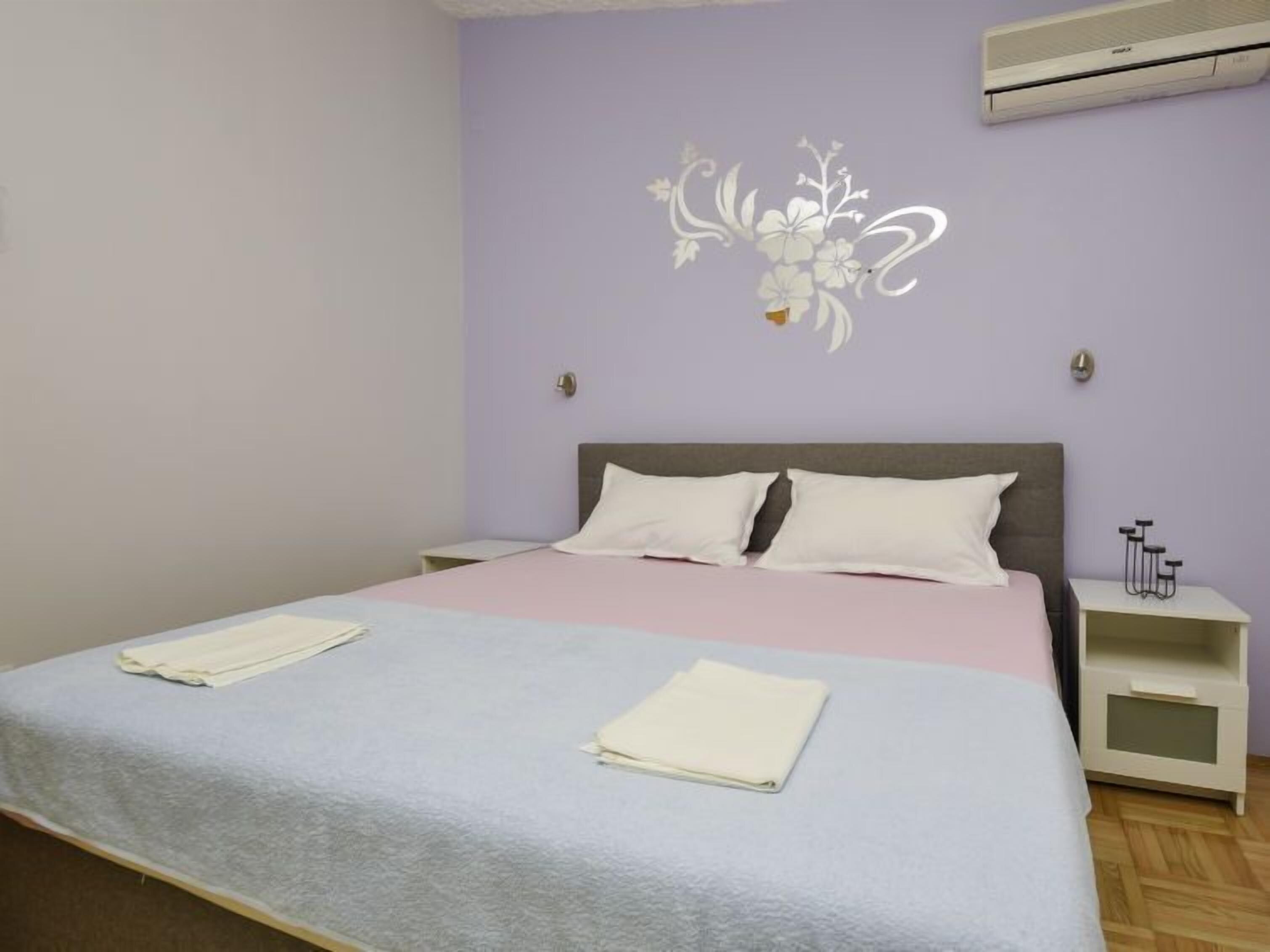 Una cama bien tendida con edredón gris y blanco, una mesita de noche con lámpara y un aire acondicionado de pared.