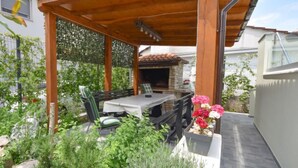 Eine überdachte Terrasse mit Tisch und Stühlen, umgeben von Grünpflanzen und einem Kamin aus Stein.