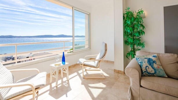 Un balcón amplio con vista al mar, un sofá blanco, una silla y una mesita con un jarrón azul.