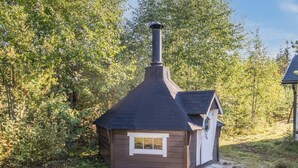Una pequeña cabaña de madera con chimenea, rodeada de árboles frondosos y un cielo despejado.
