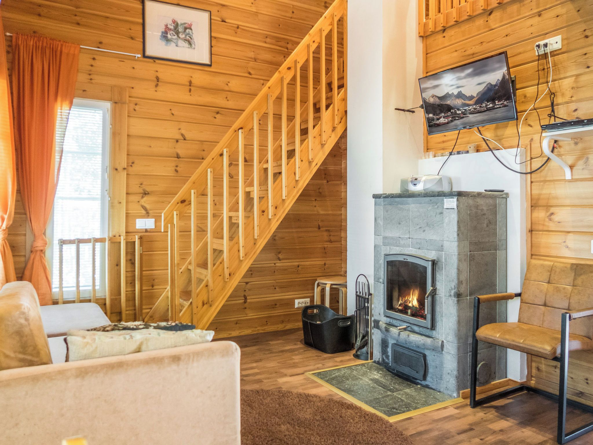 A cozy cabin interior with a wooden staircase, a fireplace, a sofa, and a television mounted on the wall.