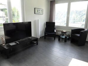 A modern living room with a large flat-screen TV, two black armchairs, a glass coffee table, and a framed picture on the wall.
