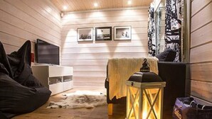 A cozy room with wooden walls, a white sofa, a black bean bag, a small table with a lantern, and a TV on a white cabinet.