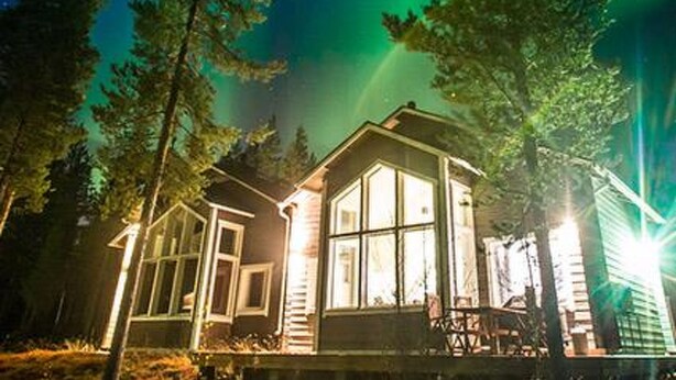 A cabin with large windows, surrounded by trees, under a night sky with visible aurora borealis.
